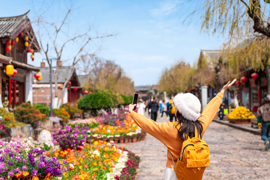 Mujer con mochila amarilla y gorro blanco tomándose una selfie con los brazos abiertos en una calle floreada del casco antiguo de Yunnan, China.